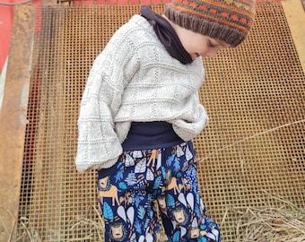 May include: A child wearing a brown and orange knit hat with a white sweater and blue pants with a jungle animal print.