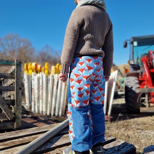 Op de afbeelding: Een kind draagt een bruine trui, een zwarte muts en een blauwe spijkerbroek met een vossenprint. Het kind staat op een houten platform.