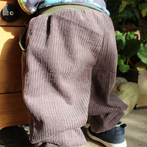 May include: A child wearing brown corduroy trousers with a green waistband and a blue and white patterned shirt. The shirt has the word "Boosch" on the side.