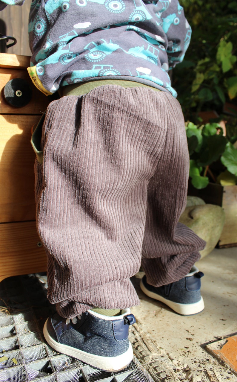 May include: A child wearing brown corduroy trousers with a green waistband and a blue and white patterned shirt. The shirt has the word "Boosch" on the side.