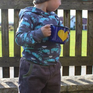 May include: A young child wearing a grey and blue knitted hat and a grey hoodie with a blue and white truck pattern holds a blue enamel mug with a yellow heart on it. The child is standing in front of a wooden fence.