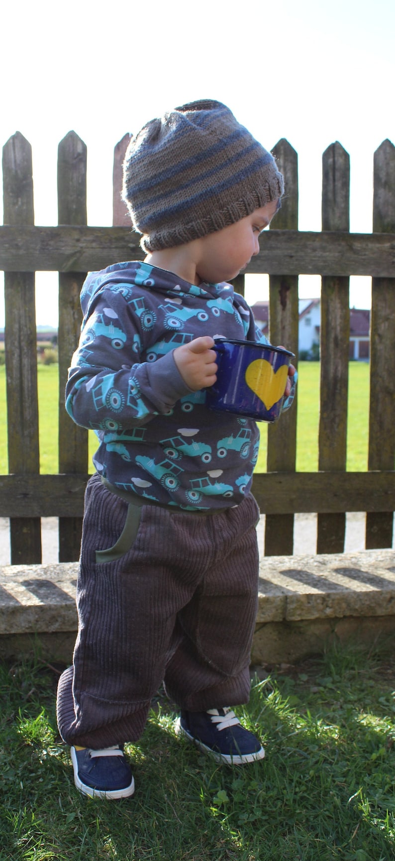 May include: A young child wearing a grey and blue knitted hat and a grey hoodie with a blue and white truck pattern holds a blue enamel mug with a yellow heart on it. The child is standing in front of a wooden fence.