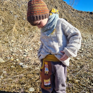 May include: A child wearing a brown and orange striped knit hat, a light grey sweater, and brown corduroy pants with a yellow waistband and a pattern of cartoon figures wearing crowns. The child is standing on a rocky, brown surface.