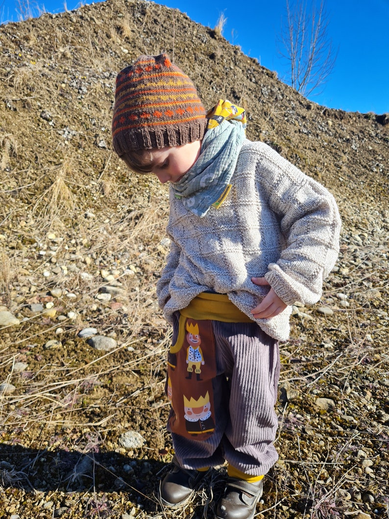 May include: A child wearing a brown and orange striped knit hat, a light grey sweater, and brown corduroy pants with a yellow waistband and a pattern of cartoon figures wearing crowns. The child is standing on a rocky, brown surface.