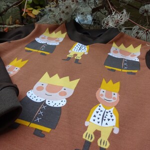 May include: Brown long-sleeved shirt with a repeating pattern of cartoon kings wearing yellow crowns and black robes. The shirt has brown sleeves.