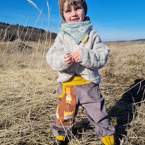 May include: A young child wearing a brown knit hat, a light green patterned scarf, a white sweater, and brown corduroy pants with a yellow waistband and a brown patch featuring a cartoon design. The child is standing in a field of tall, dry grass.