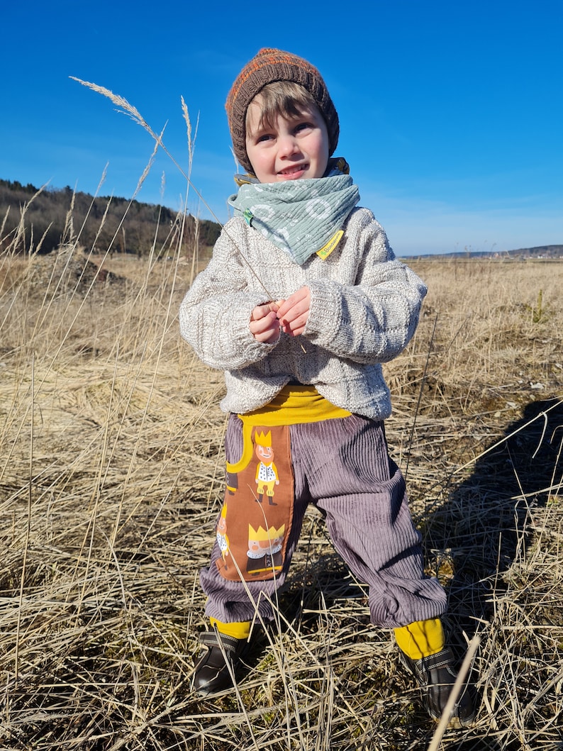 May include: A young child wearing a brown knit hat, a light green patterned scarf, a white sweater, and brown corduroy pants with a yellow waistband and a brown patch featuring a cartoon design. The child is standing in a field of tall, dry grass.