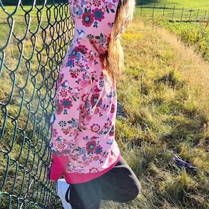 May include: A pink hoodie with a floral and bird print. The hoodie has a hood and a zipper closure. The person is wearing black pants and white sneakers.