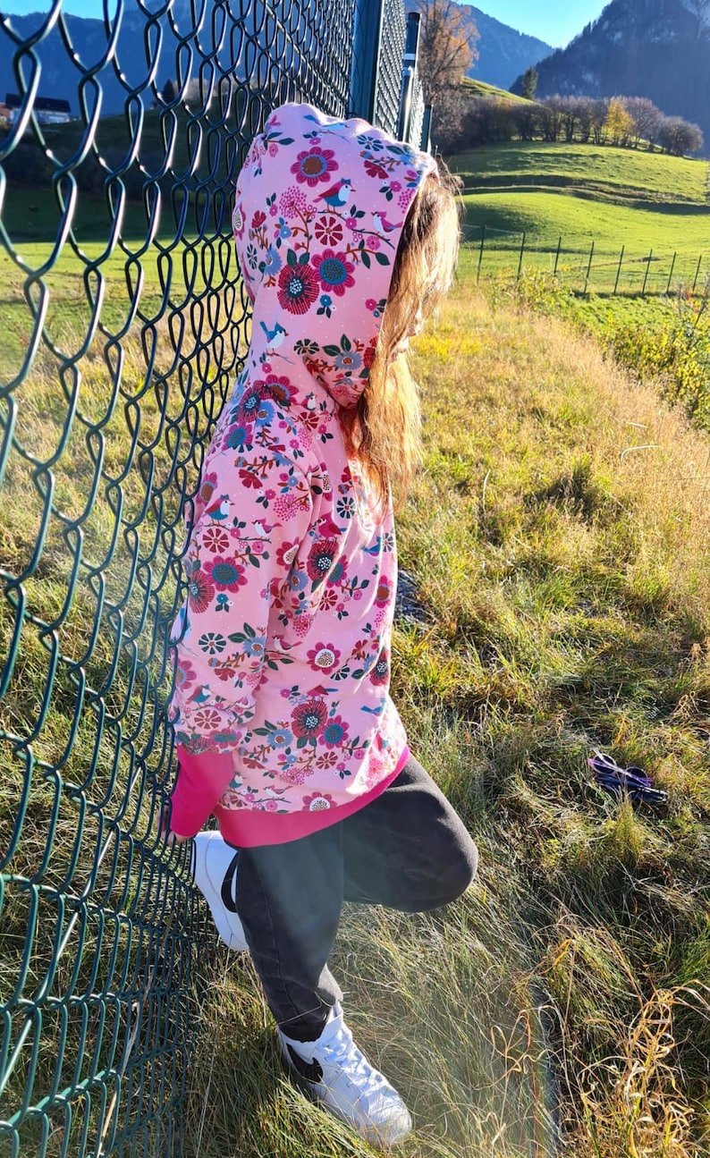 May include: A pink hoodie with a floral and bird print. The hoodie has a hood and a zipper closure. The person is wearing black pants and white sneakers.