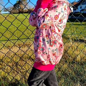 May include: A young person wearing a pink floral hoodie with a pink and black trim, black pants, and white sneakers with a black swoosh. The person is standing in front of a chain link fence.