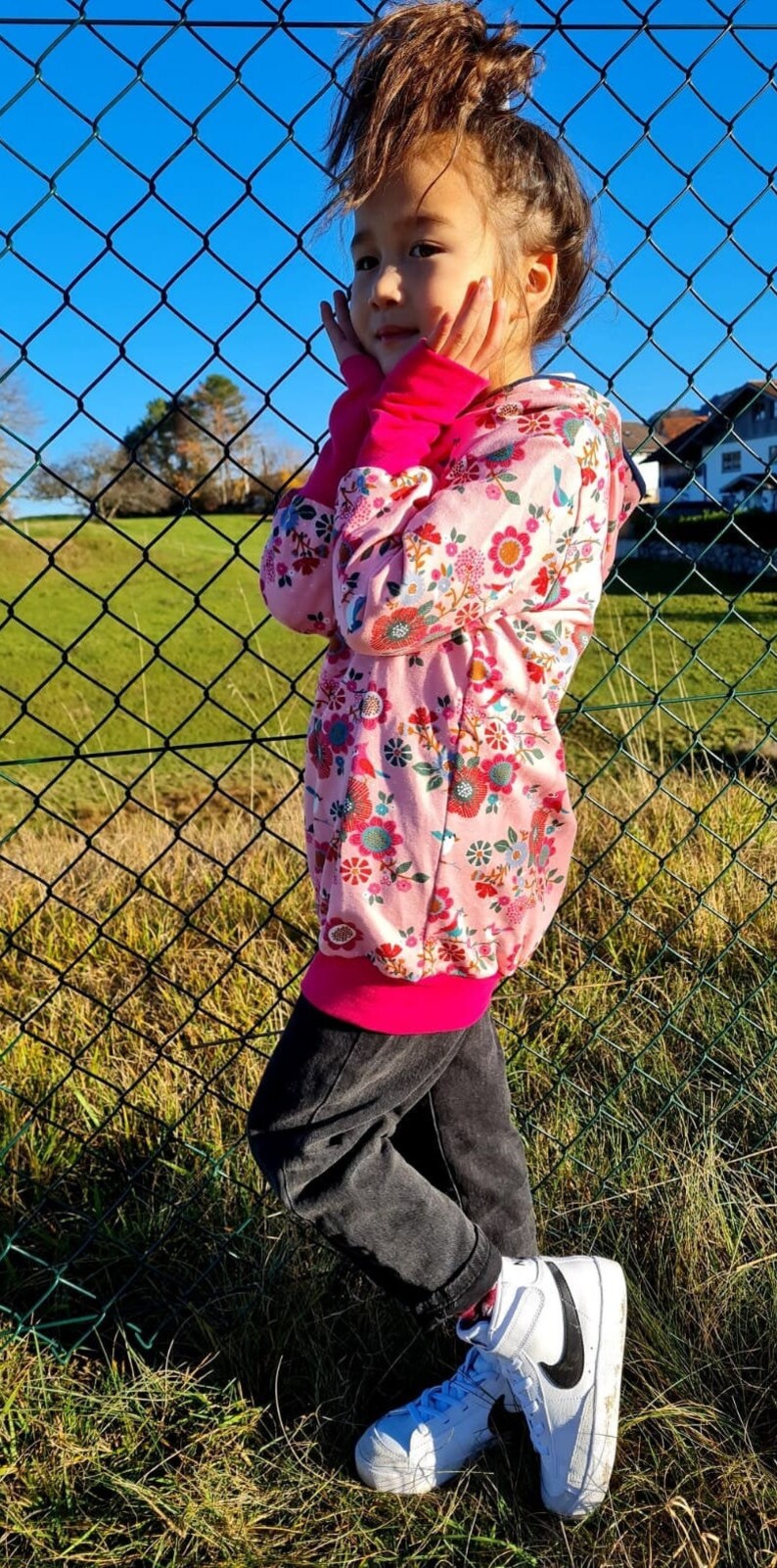 May include: A young person wearing a pink floral hoodie with a pink and black trim, black pants, and white sneakers with a black swoosh. The person is standing in front of a chain link fence.