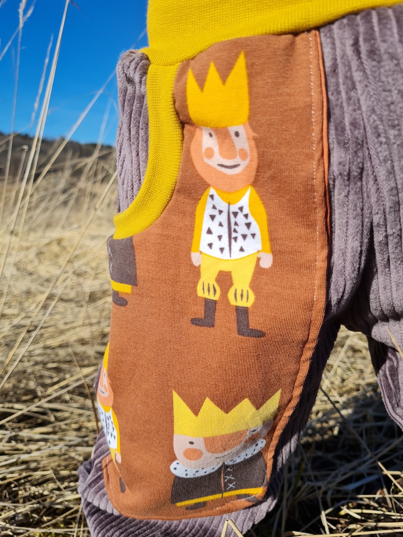 May include: Brown corduroy pants with a yellow waistband and a pattern of cartoon kings wearing gold crowns. The fabric has a textured, corduroy look.