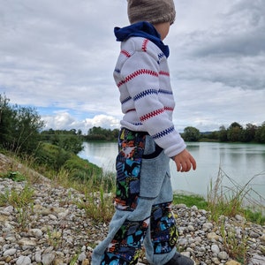 May include: A young child wearing a white and red striped jumper, a brown knitted hat, and blue jeans with a dinosaur print. The child is standing on a stony path with a body of water in the background.