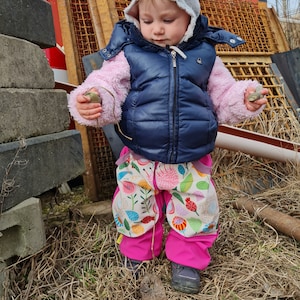 May include: A young child wearing a pink and white patterned jacket, a blue vest, and pink snow pants with a floral pattern. The child is holding two small stones in their hands and is standing in front of a red and grey metal structure.