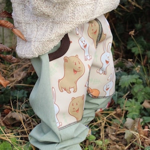 May include: A child wearing a light brown knitted sweater and green pants with a brown bear and white rabbit print. The pants have a brown and white trim.