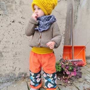 Op de afbeelding: Een kind draagt een mosterdgele kabelgebreide muts met een bijpassende pompon, een grijze sjaal en een bruin vest. Het kind draagt ook een oranje corduroy broek met bloemenpatroon manchetten en roze sneakers.