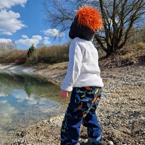 Op de afbeelding: Een kind draagt een witte trui, een donkerblauwe broek met een haaienpatroon en blauwe en gele sneakers bij een meer. Het kind draagt ook een zwarte hoed met oranje bont.