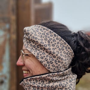 May include: A tan and black leopard print headband and neck gaiter. The ribbed fabric has a neutral background with dark spots. The neck gaiter is worn with a magenta top. The person is wearing glasses and smiling.