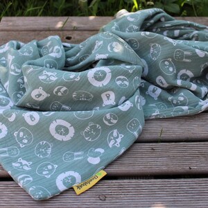 Puede incluir: Un suave arrullo de muselina verde con un estampado blanco de varios animales, incluyendo leones, tigres y mapaches. El arrullo est&aacute; doblado y tiene una etiqueta amarilla con la palabra "Buenas noches" en ella.