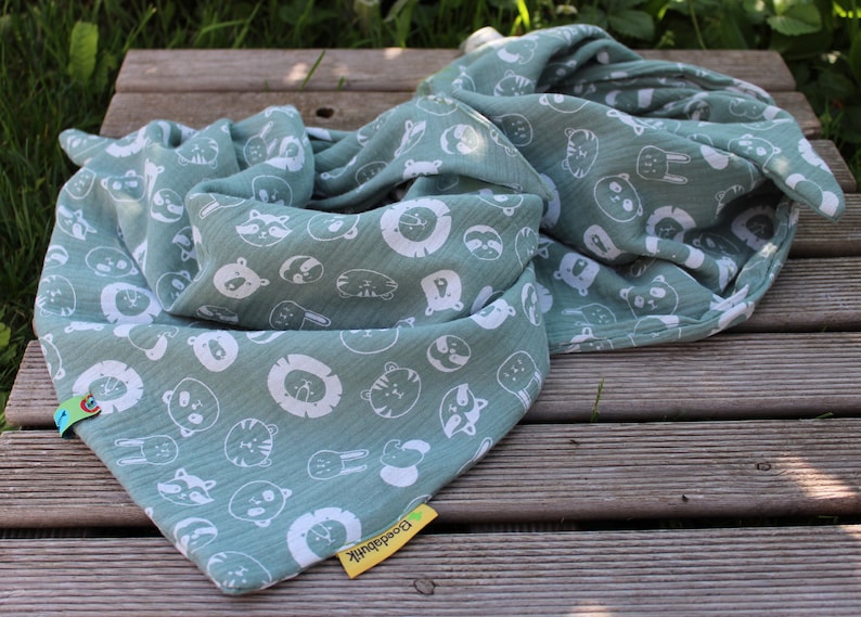 Puede incluir: Un suave arrullo de muselina verde con un estampado blanco de varios animales, incluyendo leones, tigres y mapaches. El arrullo est&aacute; doblado y tiene una etiqueta amarilla con la palabra "Buenas noches" en ella.