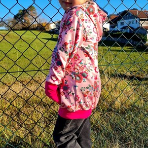 May include: A young person wearing a pink floral hoodie with a pink and black trim. The hoodie has a hood and long sleeves. The person is wearing black pants and white high-top sneakers with black accents.