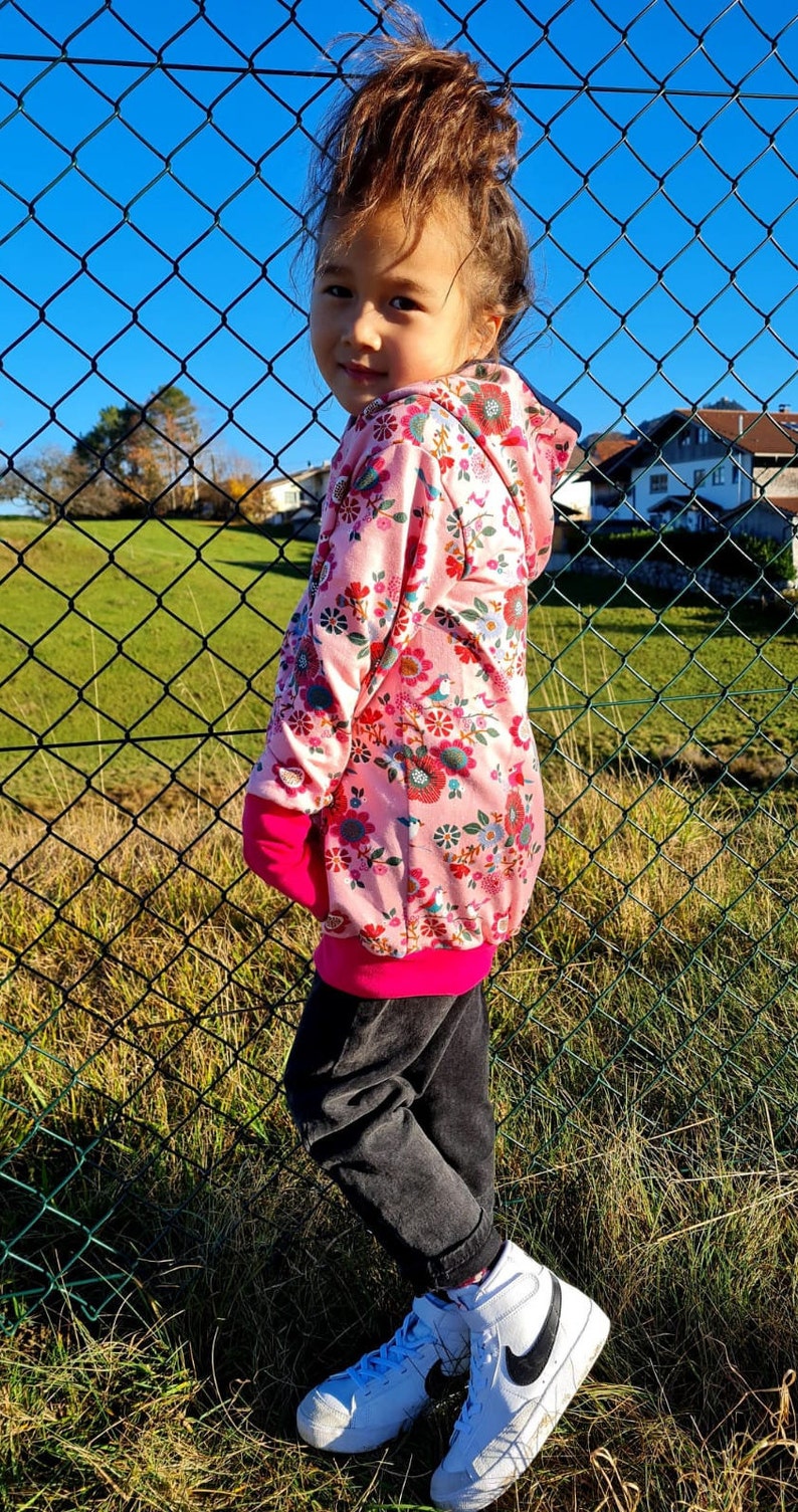 May include: A young person wearing a pink floral hoodie with a pink and black trim. The hoodie has a hood and long sleeves. The person is wearing black pants and white high-top sneakers with black accents.