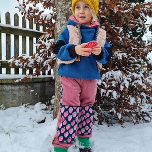 Op de afbeelding: Een jong kind draagt een blauwe hoodie, een gele gebreide muts en een roze corduroy broek met een aardbeienprint. Het kind staat in de sneeuw en houdt een rood hartvormig voorwerp vast.