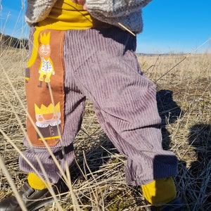 May include: Purple corduroy pants with a brown and yellow patch featuring a cartoon king. The patch has a yellow crown, a white collar, and a brown cape.