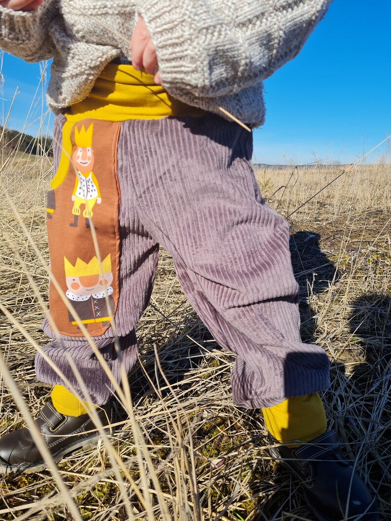 May include: Purple corduroy pants with a brown and yellow patch featuring a cartoon king. The patch has a yellow crown, a white collar, and a brown cape.