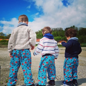 May include: Three children wearing blue pants with a red and white sea life pattern stand on a concrete surface. The children are wearing different sweaters, one white with red and blue stripes, one gray, and one blue.