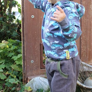 May include: A young child wearing a blue and grey striped knitted hat, a grey hooded sweatshirt with a blue and white truck pattern, and grey corduroy trousers. The child is standing in a grassy area and is pointing with their right hand.