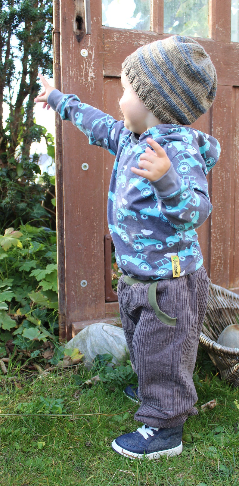 May include: A young child wearing a blue and grey striped knitted hat, a grey hooded sweatshirt with a blue and white truck pattern, and grey corduroy trousers. The child is standing in a grassy area and is pointing with their right hand.