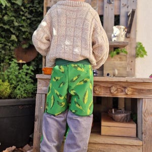 Op de afbeelding: Een kind draagt een groene pet, een beige gebreide trui en een groene korte broek met bladeren en een grijze corduroy broek. Ze dragen grijze sneakers met groene accenten, staand voor een houten speelstructuur.