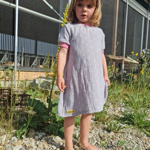 May include: A young girl wearing a light gray dress with white polka dots and a pink collar. She is standing barefoot on a gravel path with green plants in the background.