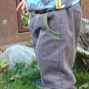 May include: A child wearing grey corduroy trousers with a green trim and a blue and grey shirt with a tractor print. The shirt has a yellow label that says "Boedabutik". The child is wearing blue trainers.