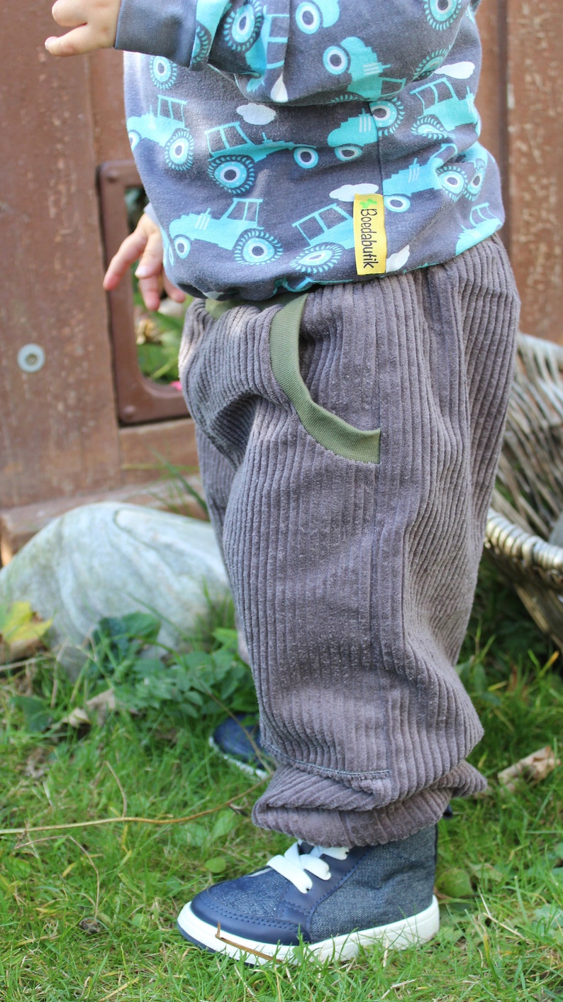 May include: A child wearing grey corduroy trousers with a green trim and a blue and grey shirt with a tractor print. The shirt has a yellow label that says "Boedabutik". The child is wearing blue trainers.