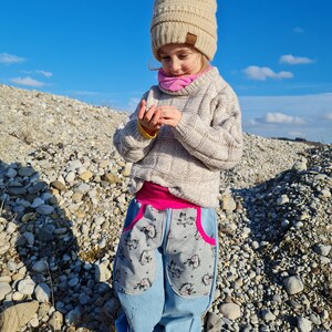 Puede incluir: Un niño pequeño con un gorro de punto beige con pompón, una bufanda rosa, un jersey gris claro y unos vaqueros azules con bolsillos estampados en gris y rosa. El niño está de pie sobre un lecho de pequeñas piedras grises.