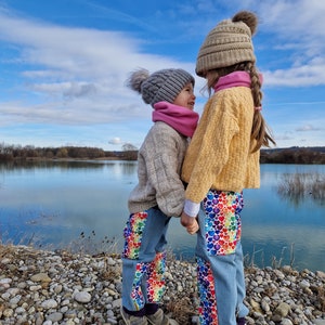 Op de afbeelding: Twee kinderen met wintermutsen en sjaals staan ​​tegenover elkaar op een stenen oever. Ze dragen allebei een blauwe jeans met kleurrijke hartpatronen. De achtergrond is een blauw meer en een bewolkte lucht.