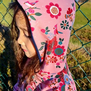 May include: A pink hooded sweatshirt with a floral and bird print. The hood is lined with a dark blue trim.