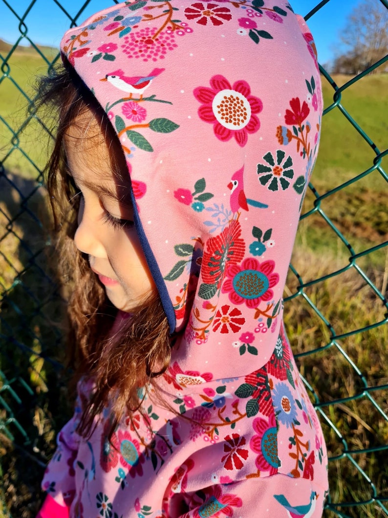 May include: A pink hooded sweatshirt with a floral and bird print. The hood is lined with a dark blue trim.