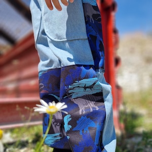 Op de afbeelding: Lichtblauwe spijkerbroek met donkerblauwe dinosaurussenprint panelen aan de zijkanten. De broek heeft een patchwork design en wordt gedragen met grijze sneakers en zwarte sokken. De foto is gemaakt op een zonnige dag buiten.