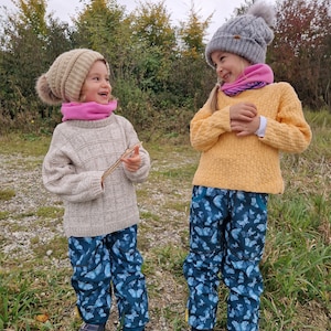 May include: Two children wearing winter hats and scarves are laughing while wearing blue pants with a butterfly pattern. The pants have pockets on the sides. The children are standing in a grassy area.