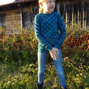 May include: A young person wearing a teal blue long-sleeved shirt with a white anchor pattern and blue jeans. They are standing in a grassy field with a wooden building in the background.