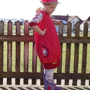 Puede incluir: Una persona joven lleva un vestido de lunares rojos y blancos con estampado floral en las mangas. Lleva un sombrero de paja con una flor rosa y botas de lluvia moradas.