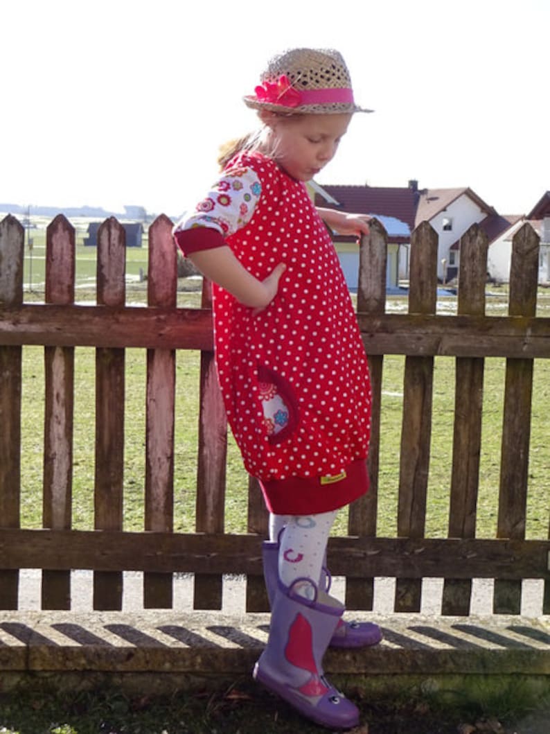Puede incluir: Una persona joven lleva un vestido de lunares rojos y blancos con estampado floral en las mangas. Lleva un sombrero de paja con una flor rosa y botas de lluvia moradas.