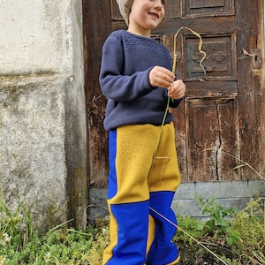 Könnte beinhalten: Ein kleines Kind trägt einen blauen Pullover, eine braune und weiße Strickmütze und eine gelbe und blaue Hose. Das Kind steht vor einer Holztür.
