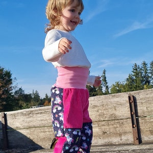 Könnte beinhalten: Ein kleines Kind trägt ein weißes Langarmshirt und eine rosa-schwarze Hose mit einem Einhorn- und Regenbogenmuster. Das Kind steht auf einer Holzoberfläche und schaut nach rechts.