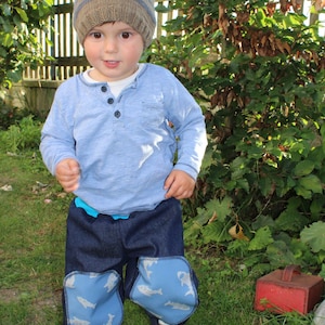 Op de afbeelding: Een jong kind draagt een blauw shirt met lange mouwen, een gestreepte gebreide muts en een blauwe spijkerbroek met een vispatroon. Het kind staat in een grasveld.
