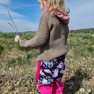 Op de afbeelding: Een kind draagt een bruine gebreide trui, een roze sjaal en een kleurrijke broek. De broek heeft een marineblauwe eenhoornprint met roze corduroy panelen. Het kind houdt een paardenbloem vast in een zonnig veld.