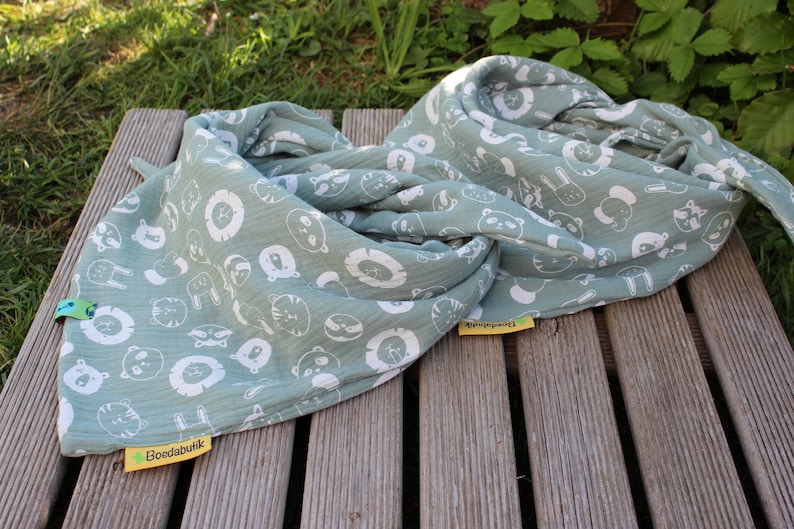 Puede incluir: Una manta de muselina verde salvia con estampado de animales blancos. La manta est&aacute; doblada y tiene una etiqueta amarilla con el texto "Boedabuik".